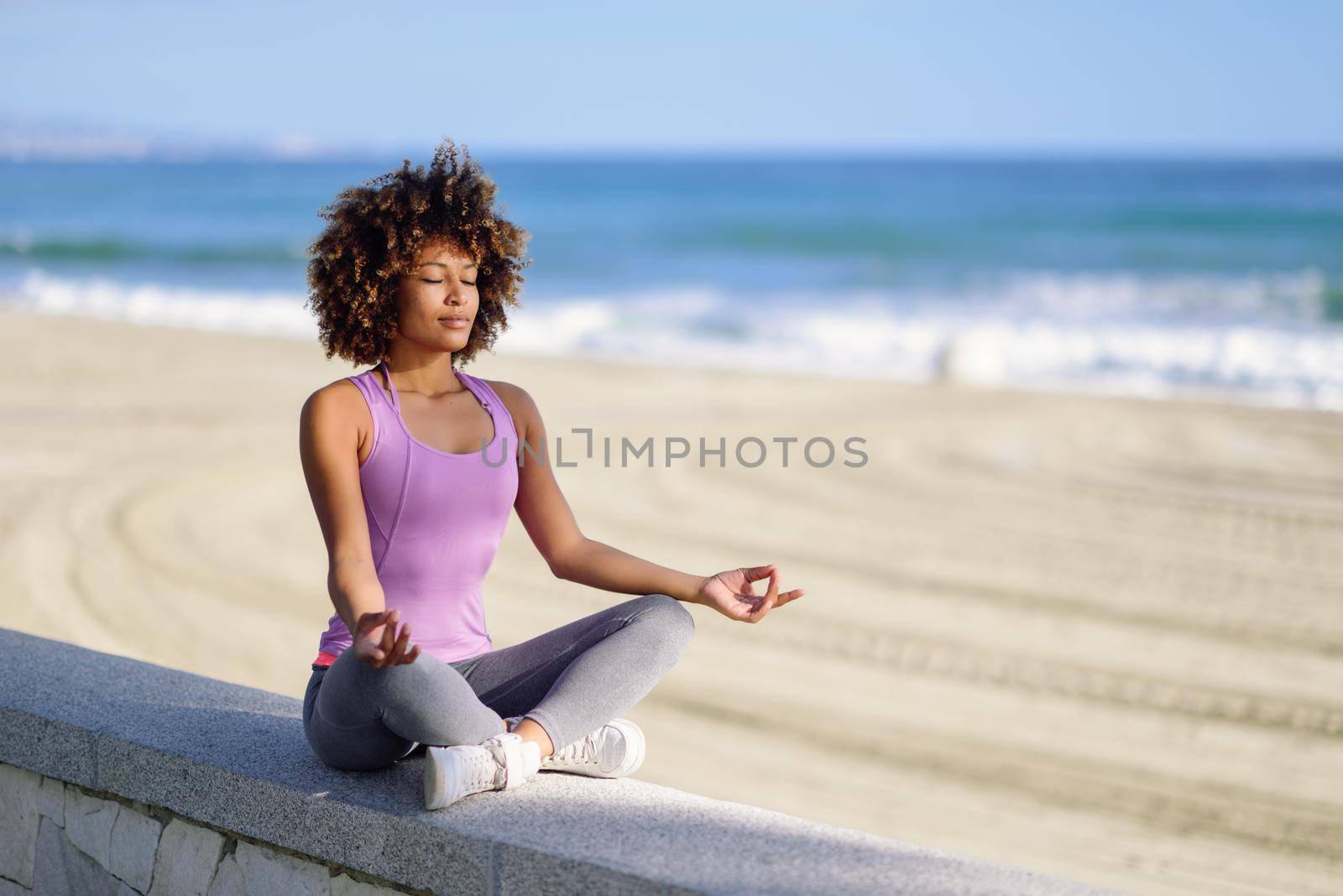 Black woman, afro hairstyle, doing yoga asana in the beach with eyes closed. Young Female wearing sport clothes in lotus pose with defocused background.
