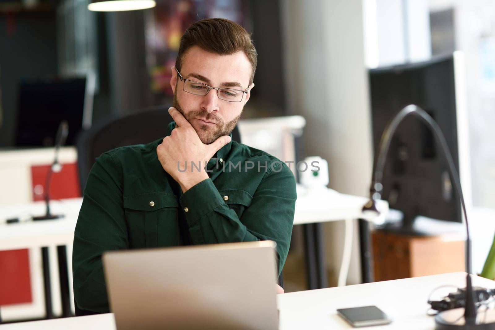 Young man studying with laptop computer on white desk. Attractive guy with beard wearing casual clothes and glasses.