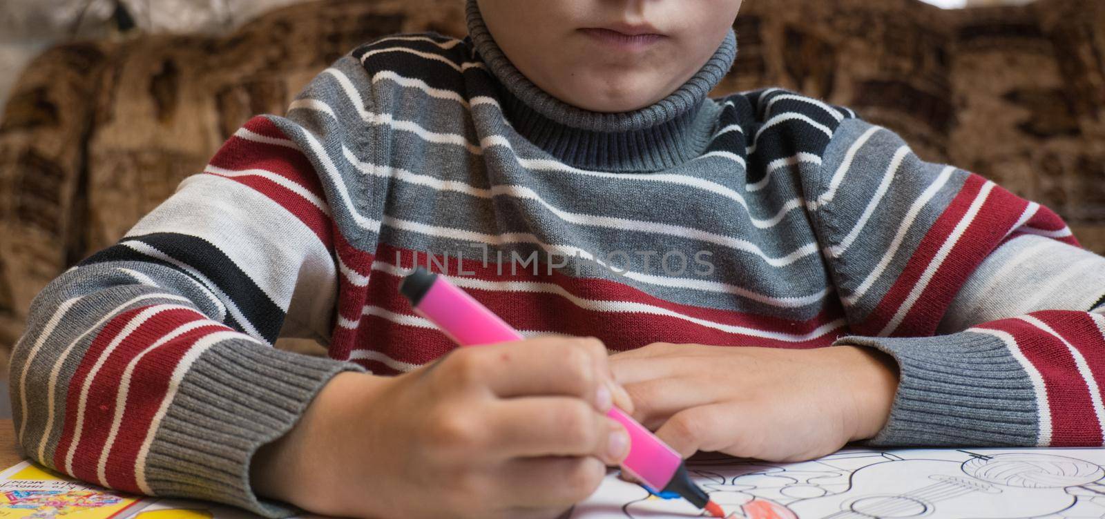A little boy paints a coloring with crayons and felt-tip pens on a wooden table at home.