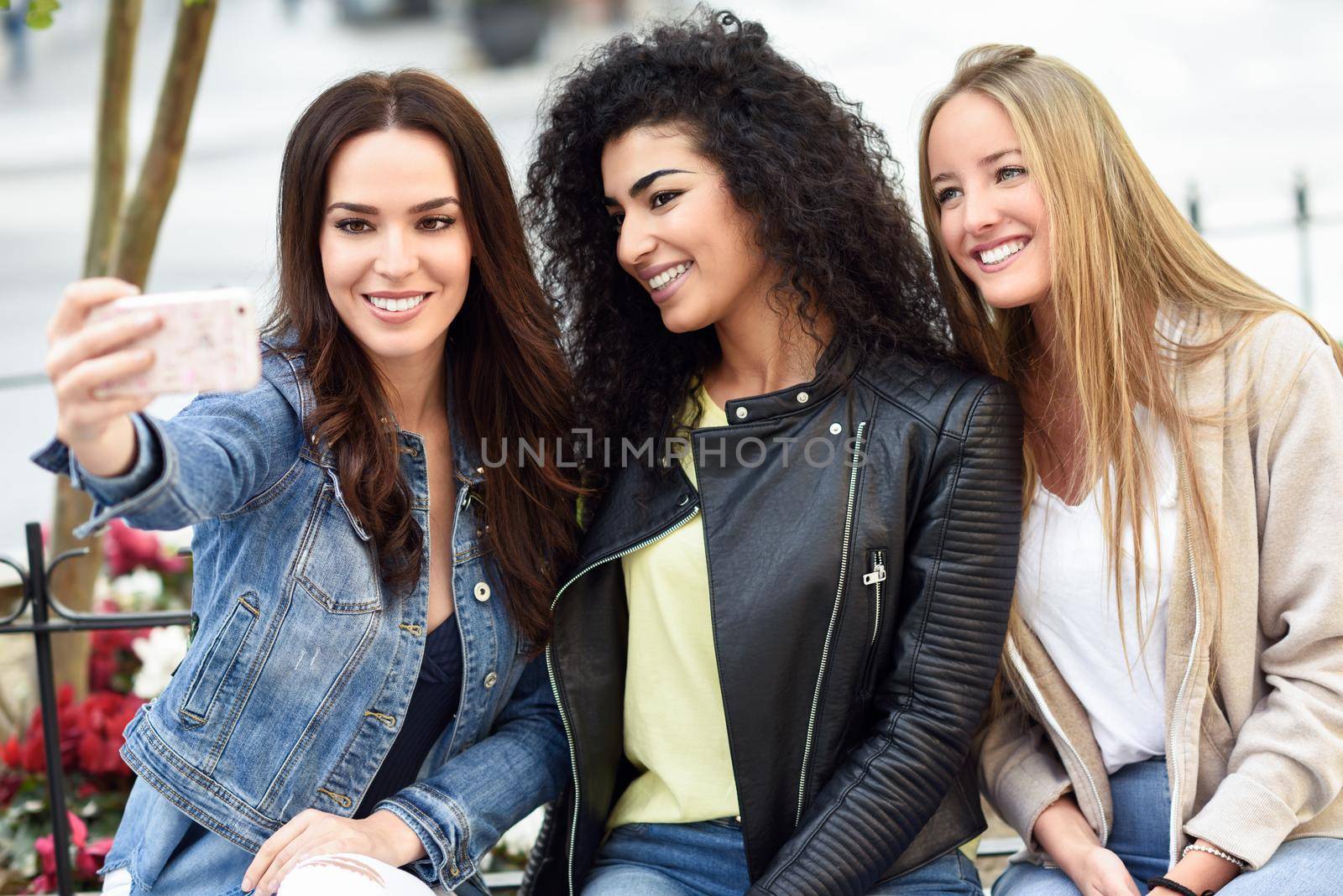 Group of multi-ethnic young women taking a selfie photograph together outdoors. Blonde, brunette and mixed females wearing casual clothes in urban background.