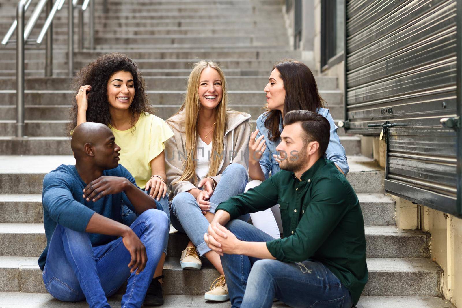 Multi-ethnic group of young people having fun together outdoors in urban background. group of men and woman sitting together on steps.