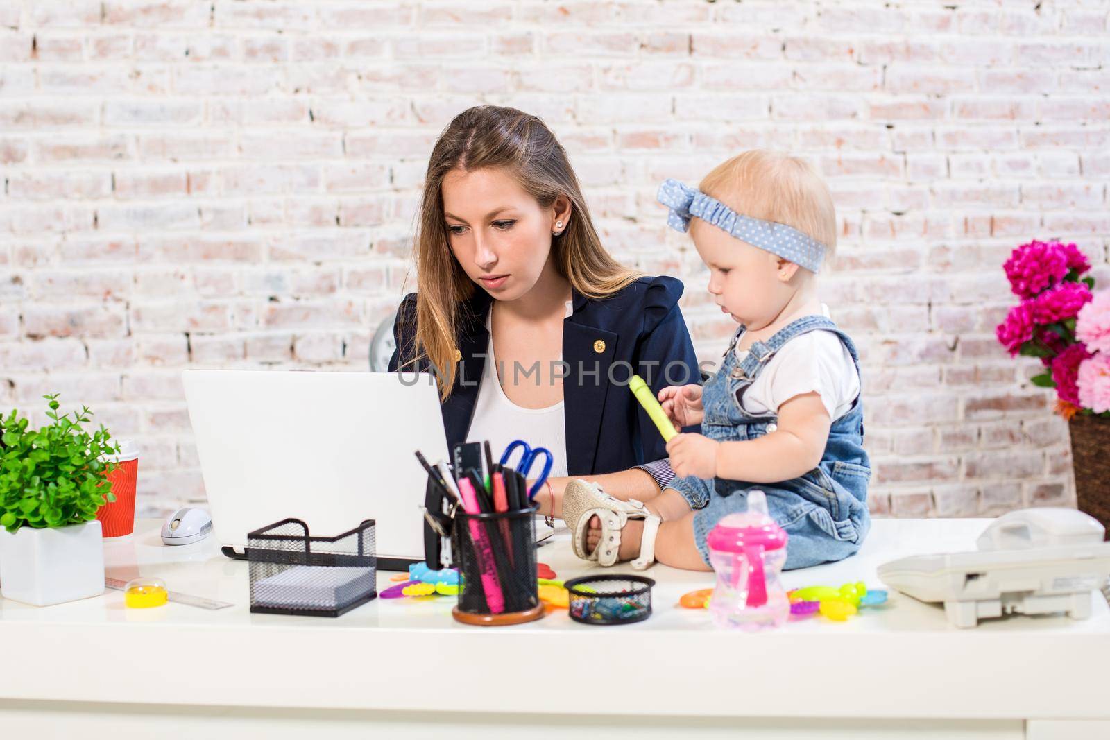 Cheerful young beautiful businesswoman looking at laptop while sitting at her working place with her little daughter by nazarovsergey