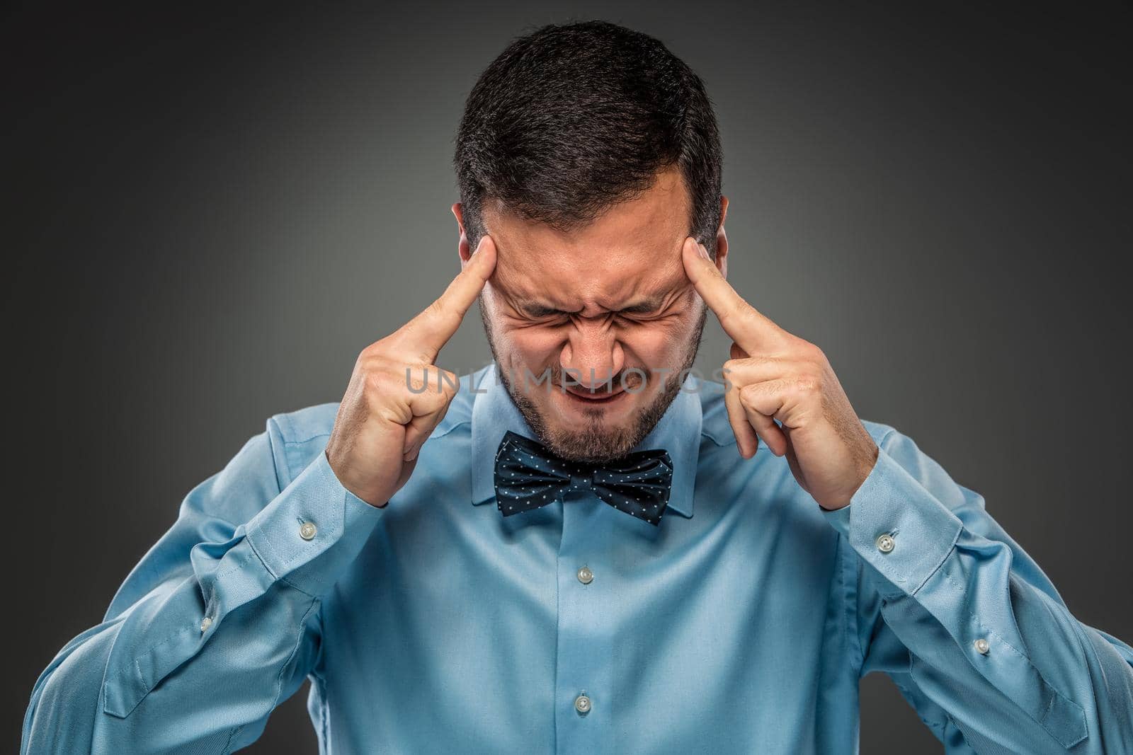Portrait of angry upset young man in blue shirt and butterfly tie, fingers touch the head, isolated on gray studio background. Negative human emotion, facial expression. Closeup