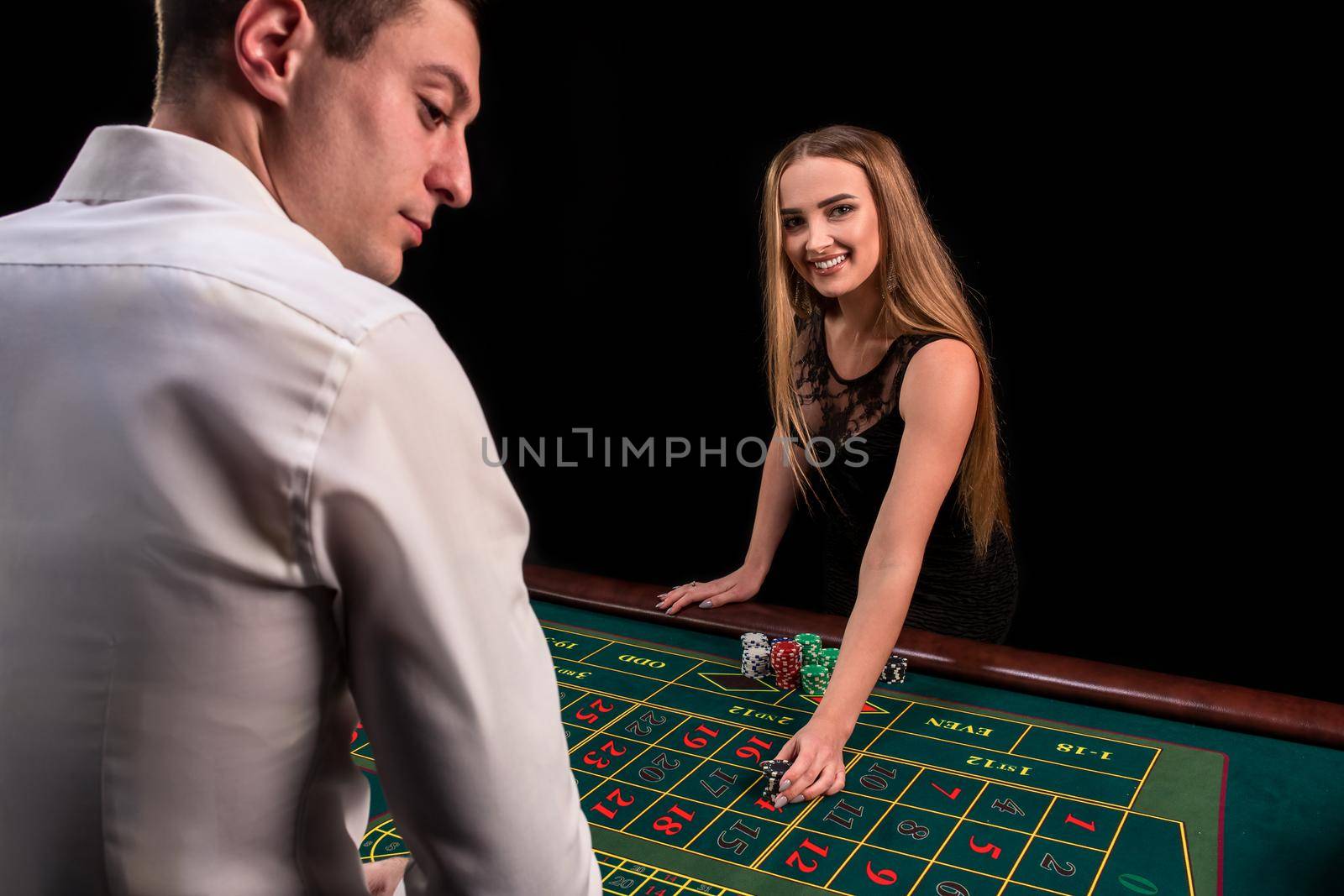 A close-up on the back of the croupier in a white shirt, image of green casino table with roulette and chips, a rich woman betting of gambling in the background. Casino. Gambling. Roulette. Betting