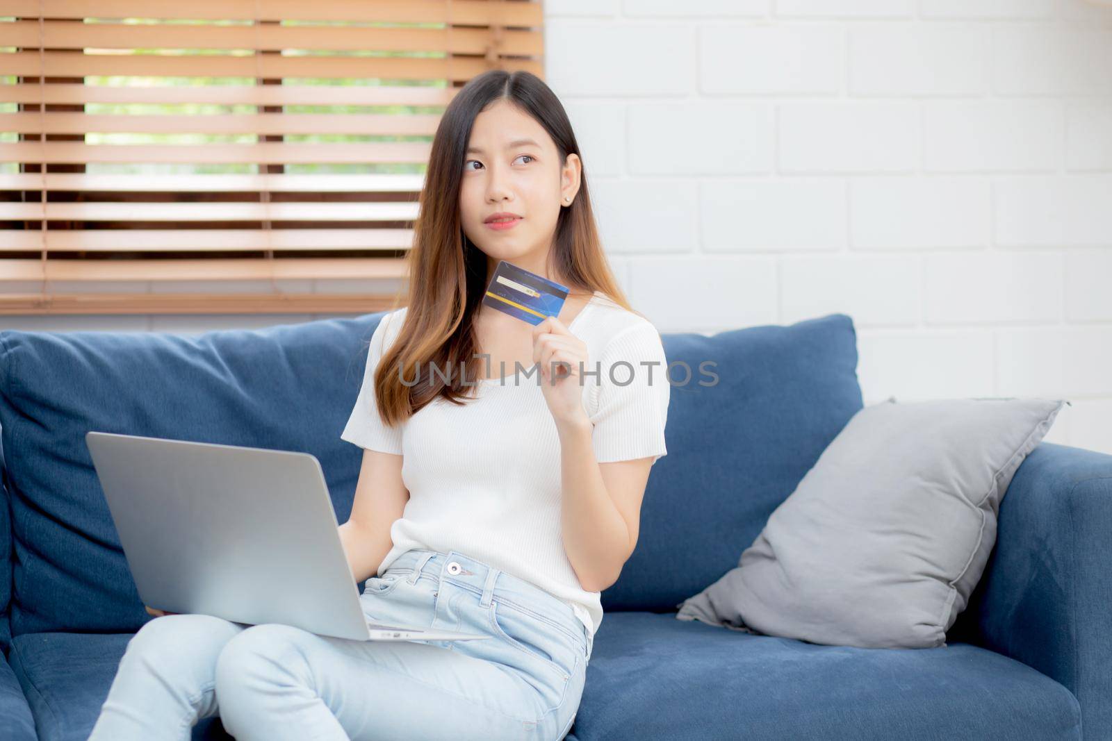 Young asian woman sitting thinking idea using credit card with laptop computer on couch, girl shopping online for buy and payment with notebook on sofa, finance and debit, lifestyle concept. by nnudoo