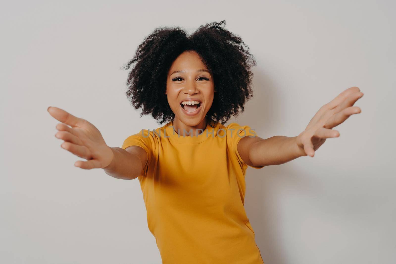 Happy to see you. Studio shot of young overjoyed african lady stretching her arms and wants to hug, smiling cheerfully at camera while posing isolated over white background, dressed in yellow tshirt