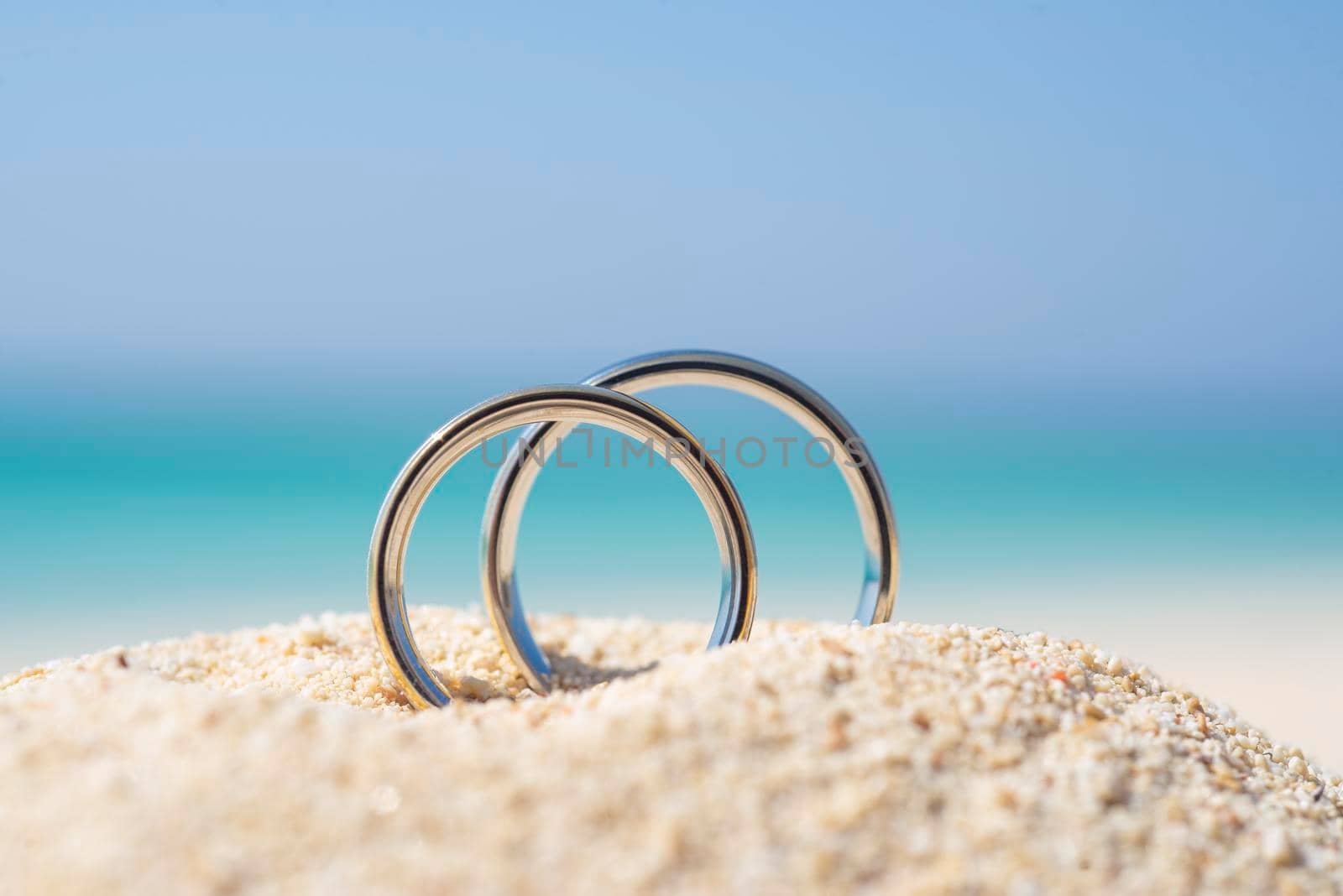 Pair of white gold wedding ring bands jewelry in sand on tropical desert island beach during summer with blue ocean background