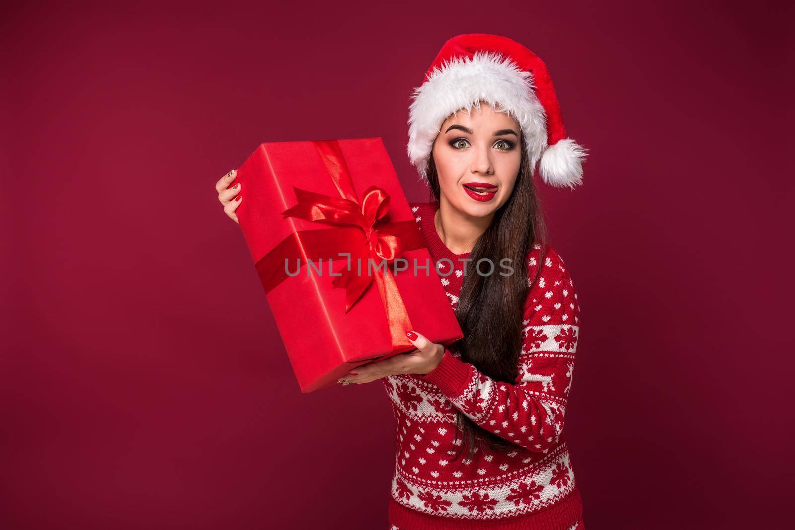 Young woman with long beautiful hair in Santa hat and New Year's dress holding Christmas present in her hands on red studio background. Christmas sale. Emotional brunette. Copy space