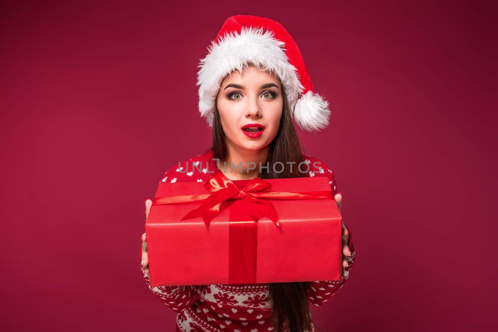 Young woman with long beautiful hair in Santa hat and New Year's dress holding Christmas present in her hands on red studio background. Christmas sale. Emotional brunette. Copy space