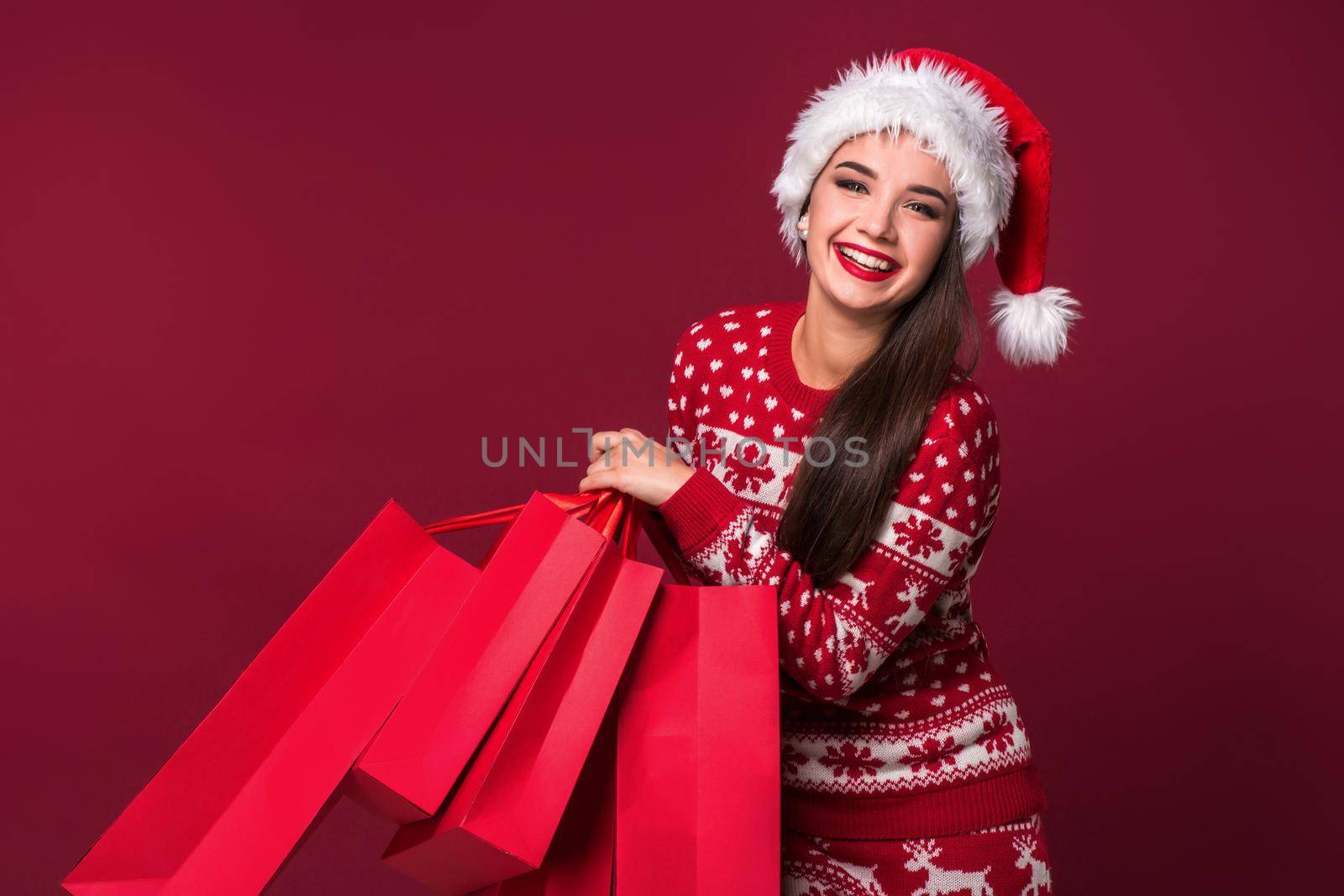 Young pretty woman or girl with long beautiful hair in Santa hat and New Year's dress holding red package on red studio background. Christmas sale. Emotional brunette. Copy space