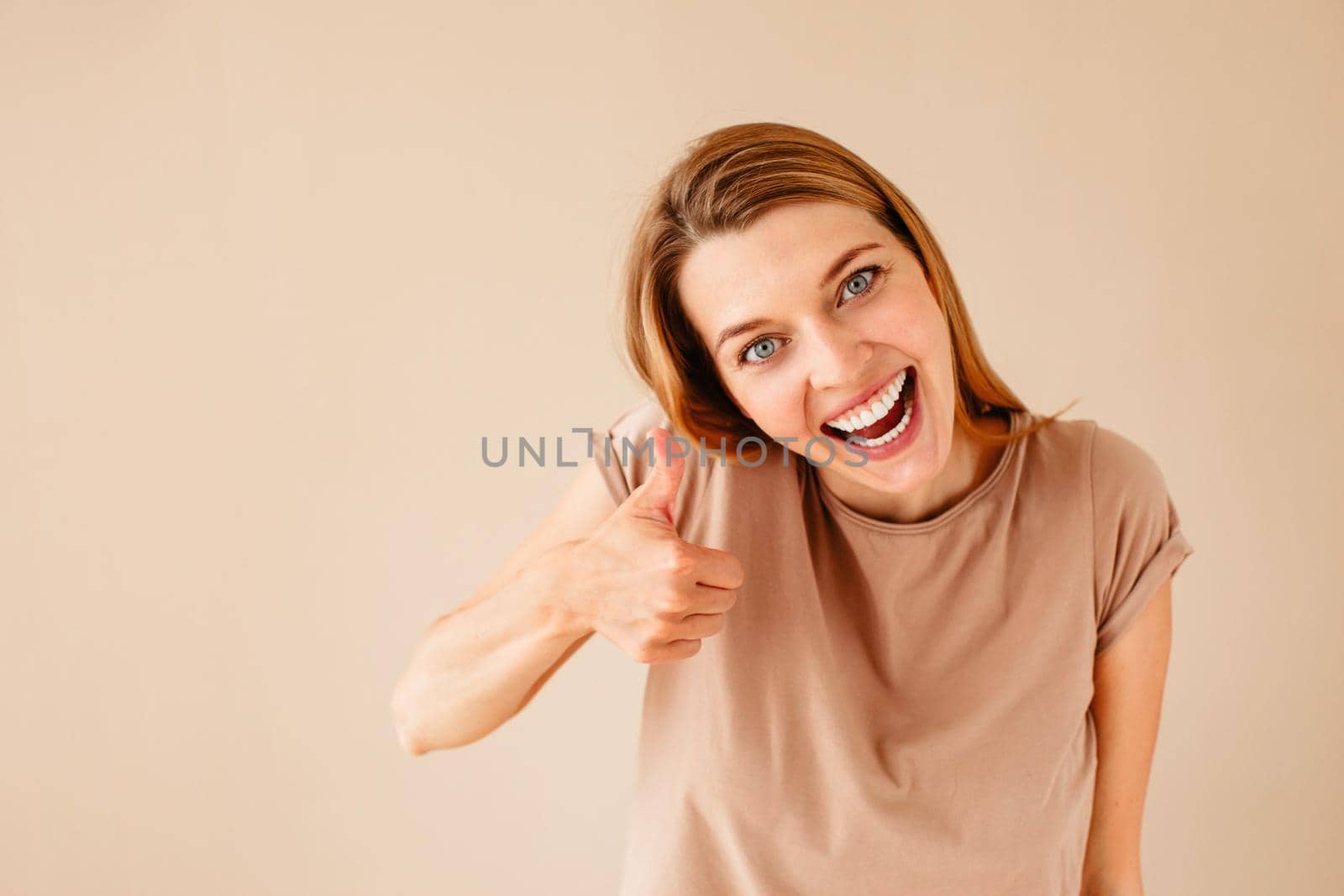 Cheerful young woman smiling and looking at camera while showing thumb up gesture against beige background