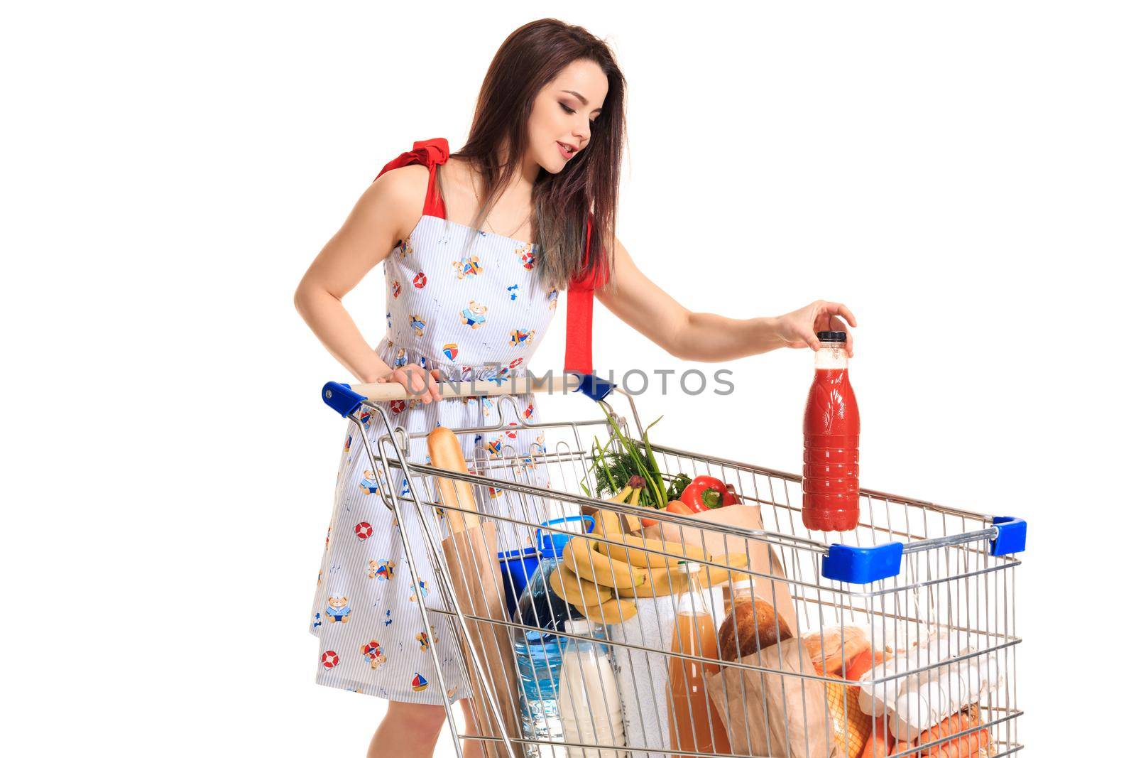 Smiling young woman doing grocery shopping at the supermarket, she is putting a tomato juice bottle in the cart isolated on white background. Brunette in a summer dress makes purchases