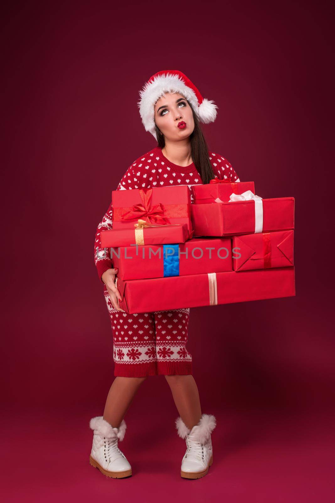 Young woman in dress and christmas hat holding gift boxes on red background. Christmas sale. Emotional brunette. Copy space