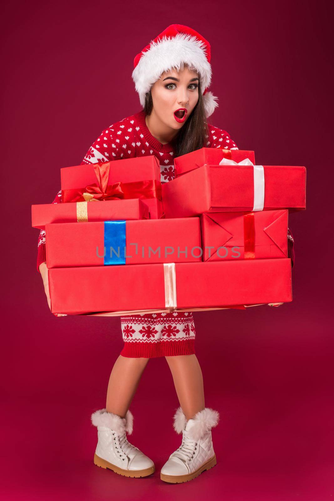 Amazed beautiful woman holding many presents on red background. Young woman in santa hat looks at camera. Christmas. Discounts. The shops. Sale