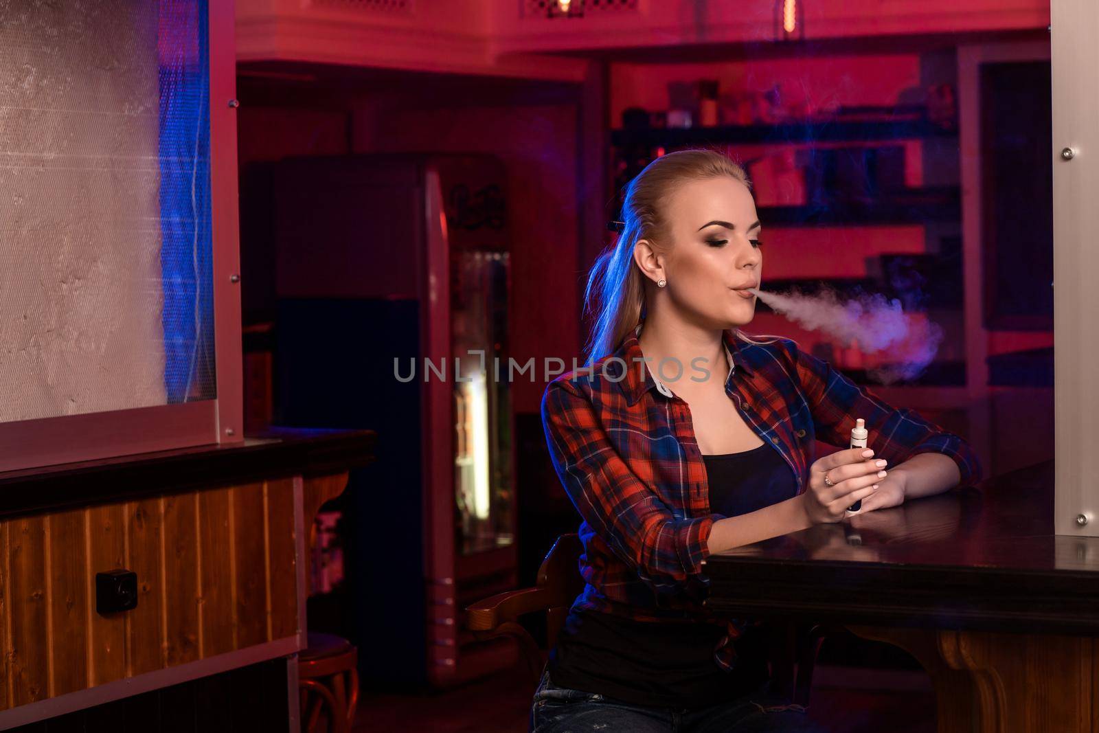 Young pretty woman in a shirt in a cage smoke an electronic cigarette at the vape bar. Vape shop.