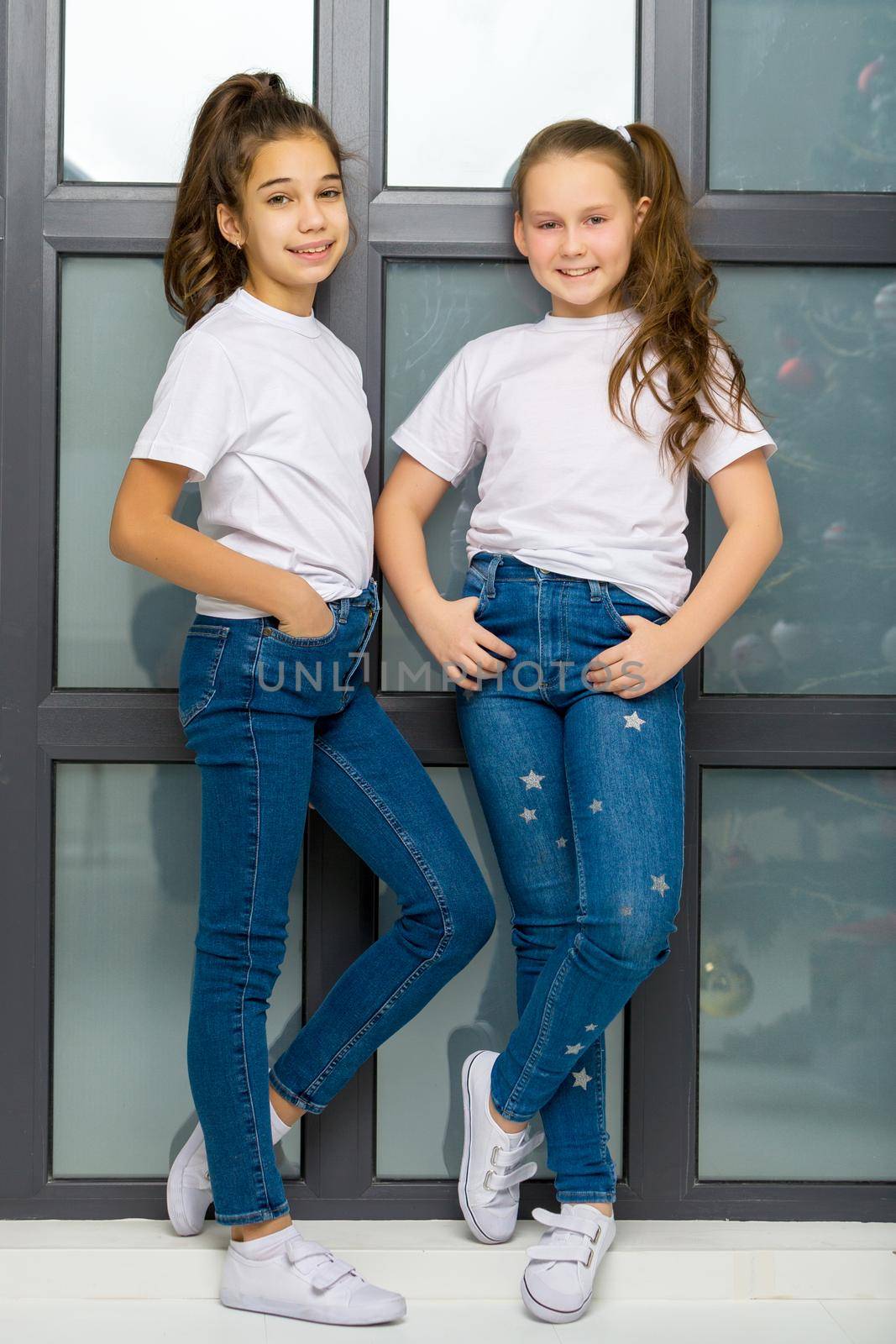 Two beautiful little girls of school age posing in the studio near a large panoramic window. Outside the window it is already evening, dark, the concept of tenderness and beauty.