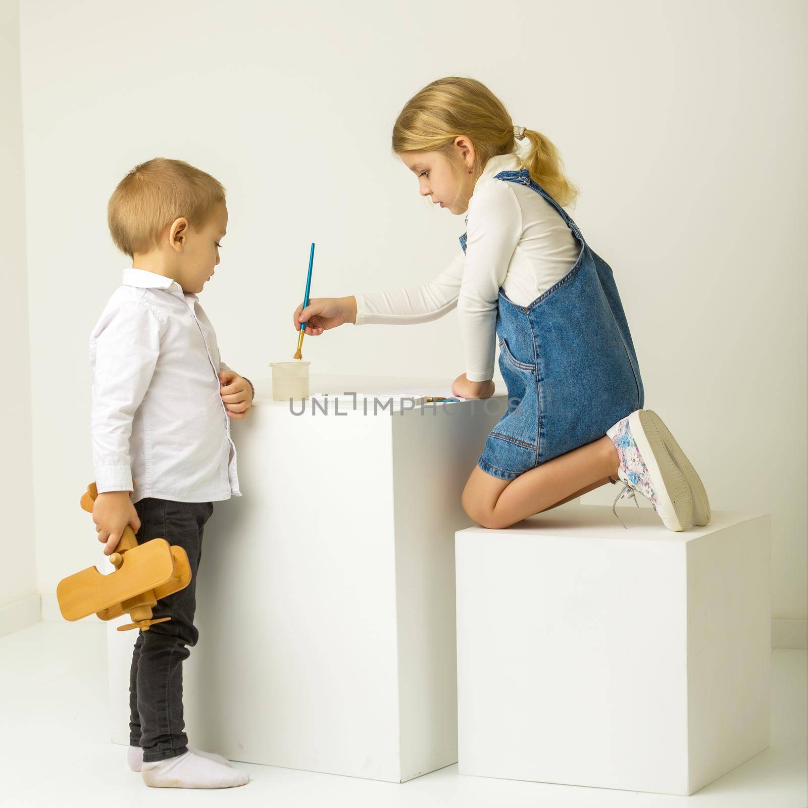 Cute Blonde Girl Sitting on Her Knees and Painting with Paintbrush, Her Brother Looking at Her with Interst, Adorable Brother and Sister in Fashionable Clothes, Portrait of Cheerful Stylish Children