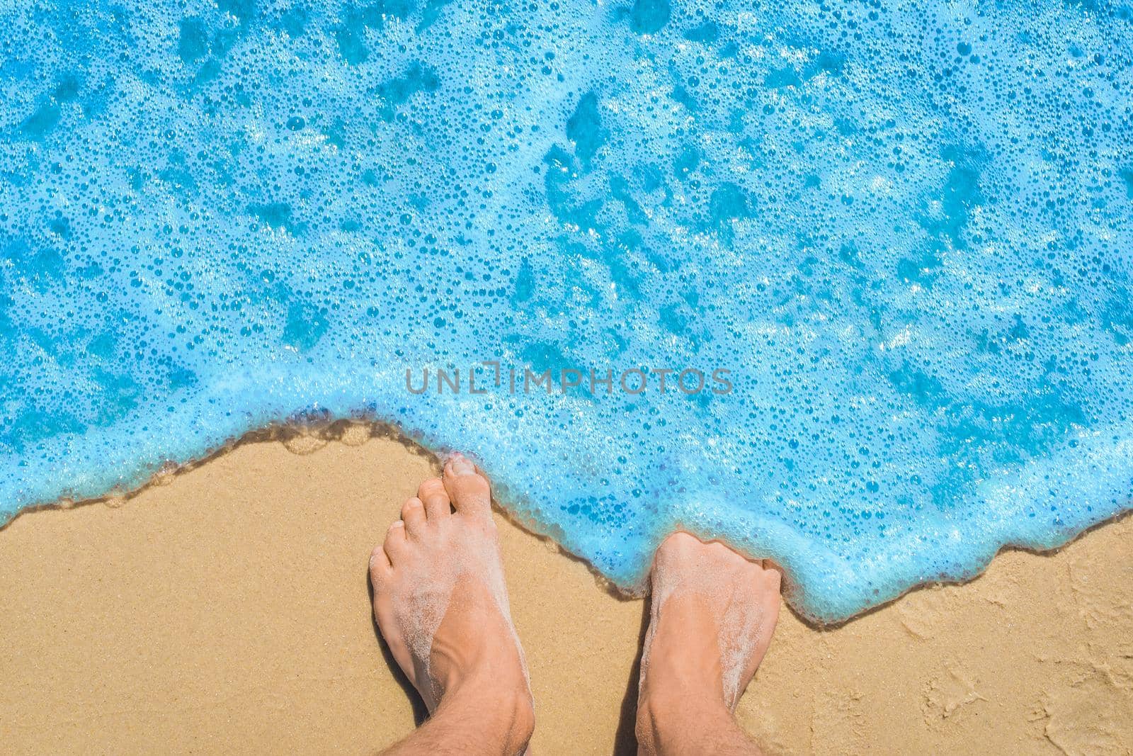 The young man's legs stand on the beach sand and are washed by blue sea water, a view from above.