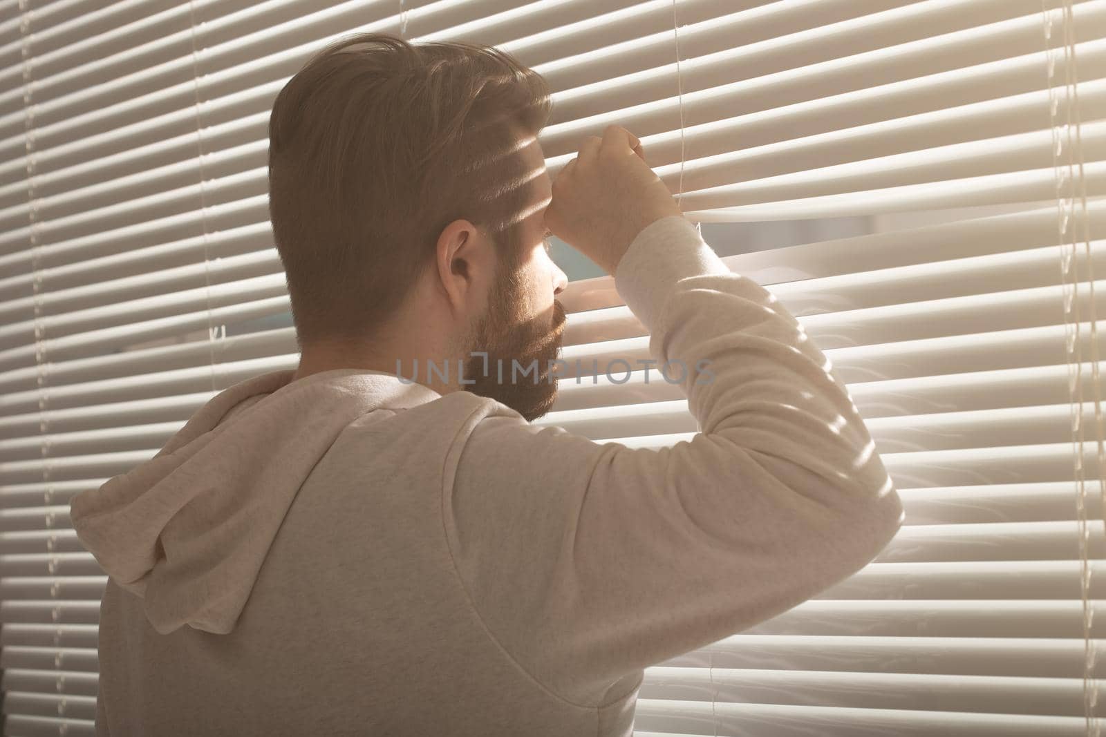 Rear view of young man with beard peeks through hole in the window blinds and looks out into the street. Surveillance and curiosity