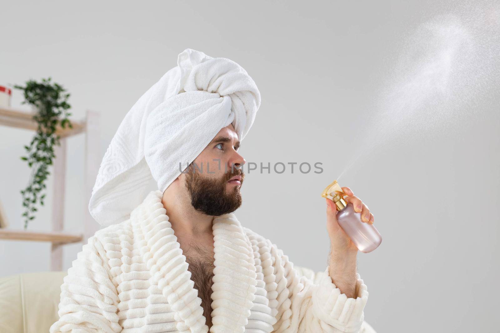 Bearded male with bath towel on his head applying spray water treatment on face. Spa, body and skin care for man concept