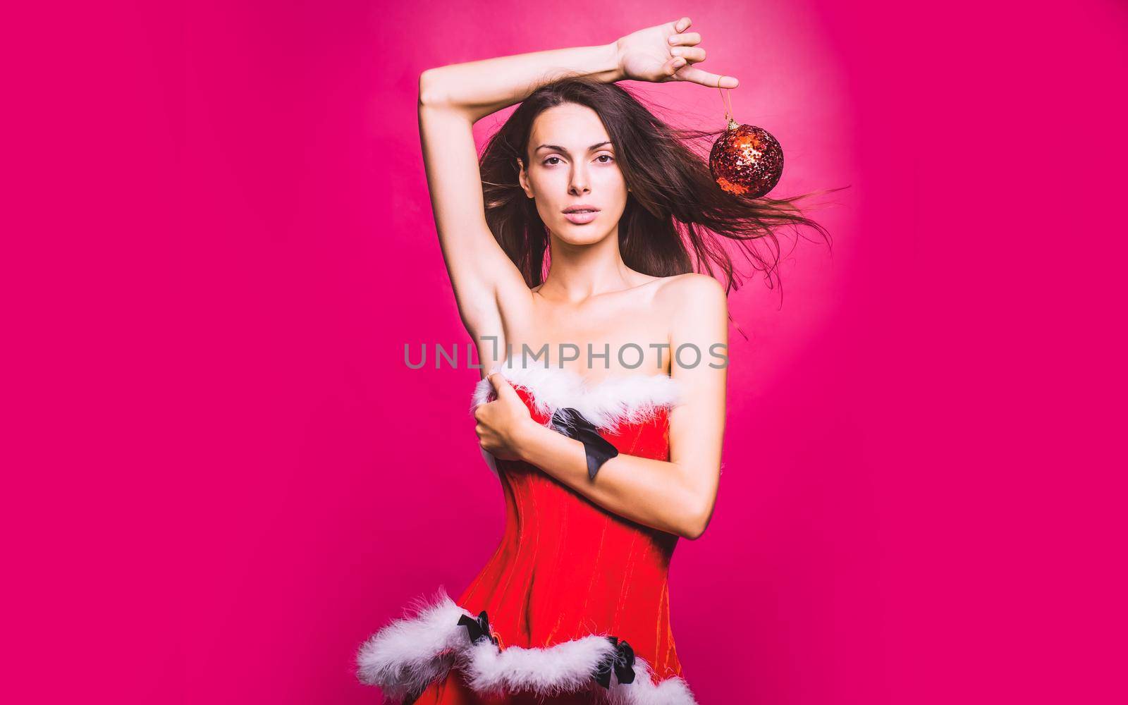 Sexy woman in red christmas santa holiday dress holds decorative ball in studio on red background.