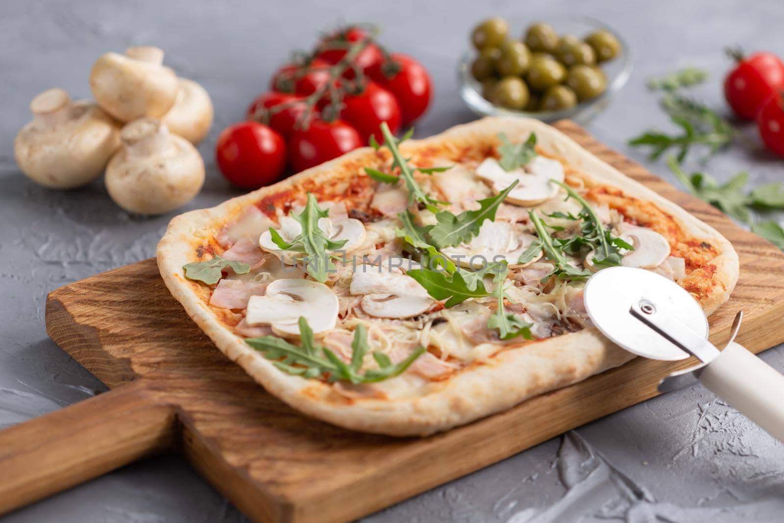 Rectangular pizza with mushrooms, tomatoes and arugula on a wooden cutting board.