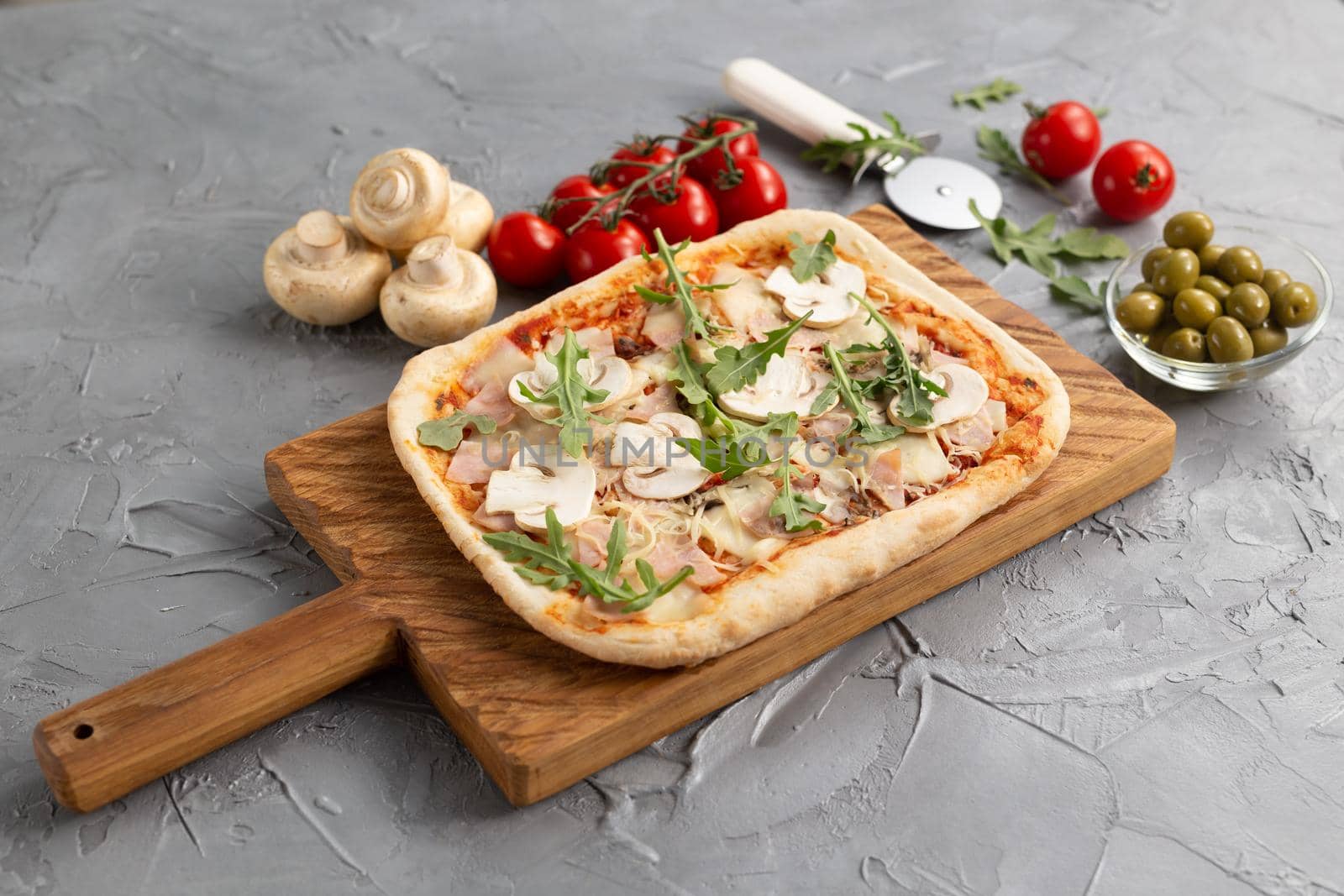 Rectangular pizza with mushrooms, tomatoes and arugula on a wooden cutting board.