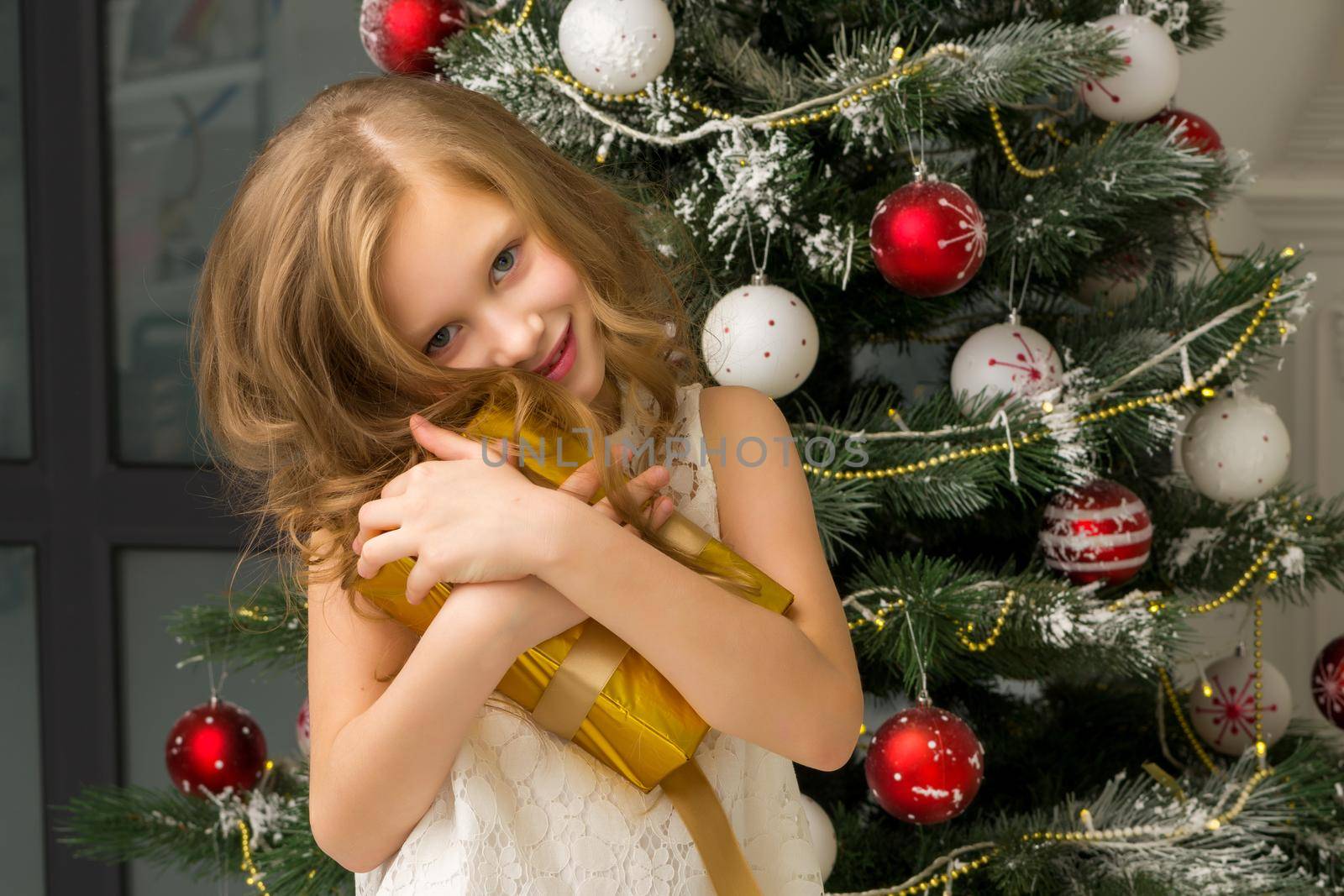 Happy Girl Standing in Front of Decorated Fir Tree Hugging Golden Gift Box, Beautiful Smiling Preteen Child Wearing Nice White Dress Posing in Christmas Interior, Lovely Girl Opening Presents on Xmas Eve