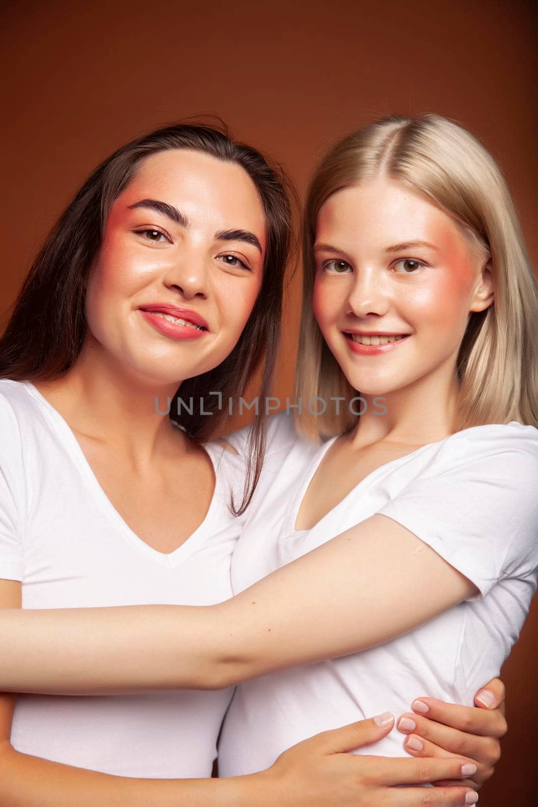 two pretty diverse girls happy posing together: blond and brunette, caucasian and asian on brown background, lifestyle people concept close up