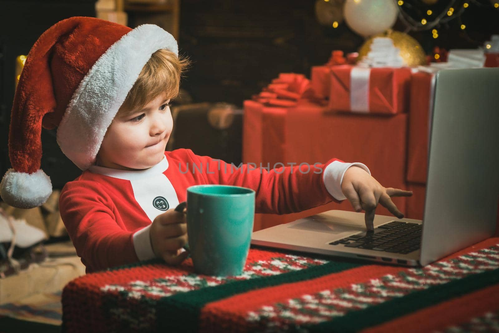 Online Christmas shopping for kids. Little child boy is wearing Santa clothes sitting by his laptop. Santa helper using notebook