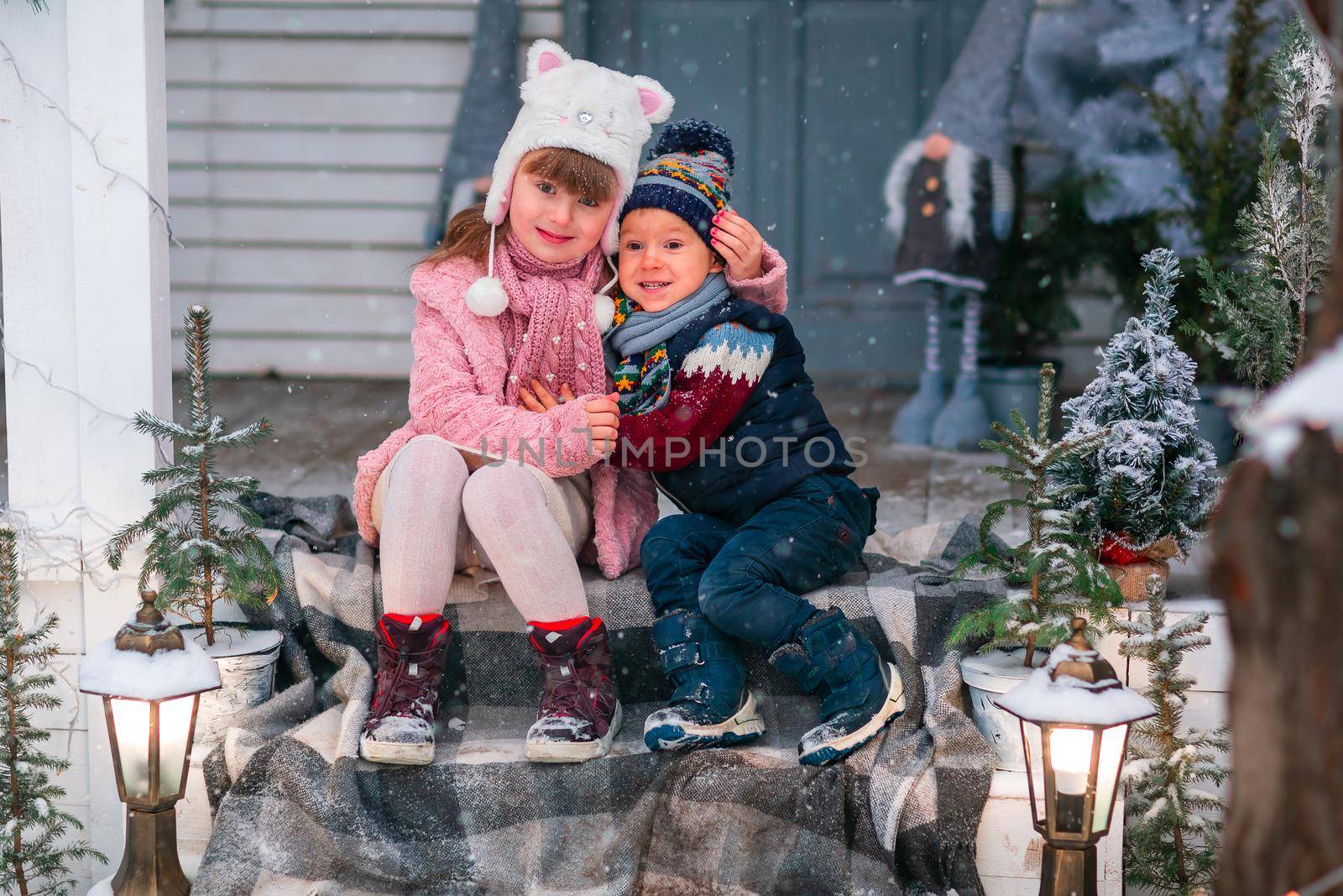 Happy little kids sitting on the porch of the Christmas decorated house, snowing outdoor. Happy New Year and Merry Christmas. Magic winter