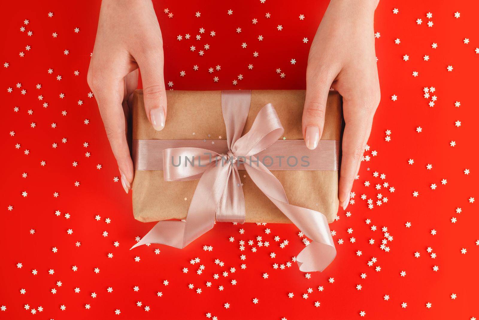Nice female hands holding gift box wrapped in kraft paper with pink ribbon on festive red background with many snowflakes. Xmas and New Year postcard design. Giving love and warmth, Christmas concept