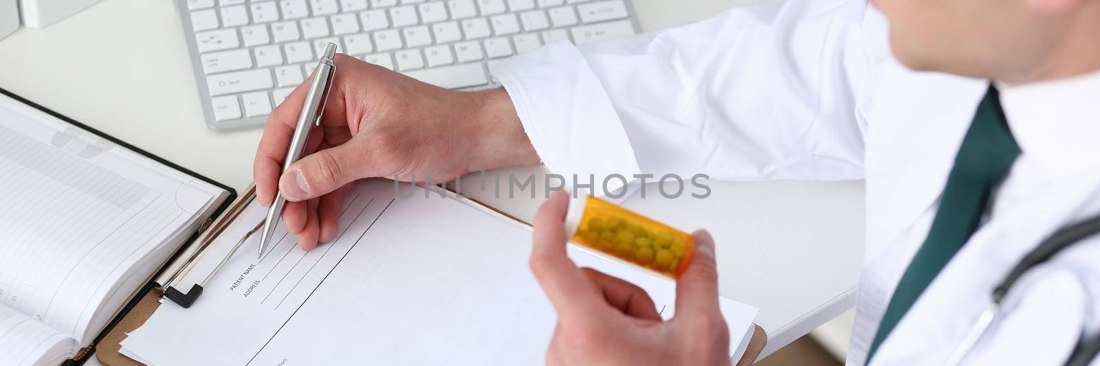 Male medicine doctor hand hold jar of pills and write prescription to patient at worktable. Panacea and life save, prescribing treatment, legal drug store concept. Empty form ready to be used