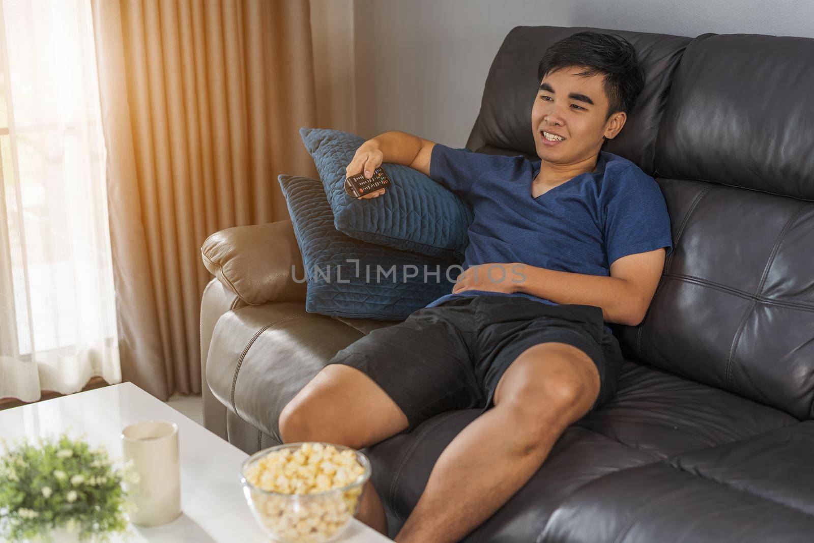 cheerful young man holding remote control and watching TV while sitting on sofa in the living room 