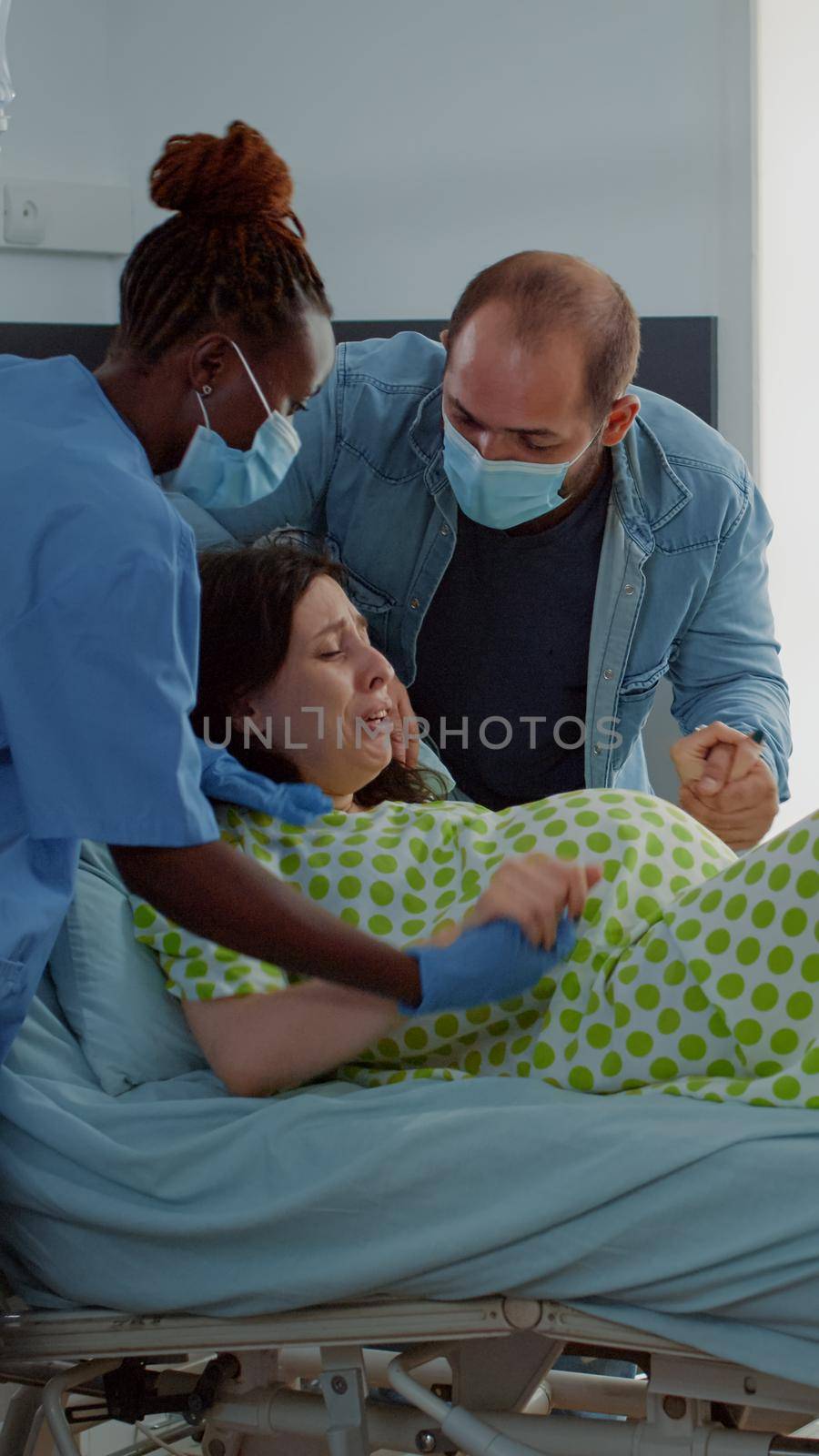 Obstetrics doctor and african american nurse assisting birth in hospital ward. Pregnant woman in labor with painful contractions pushing for baby. Young husband sitting for support