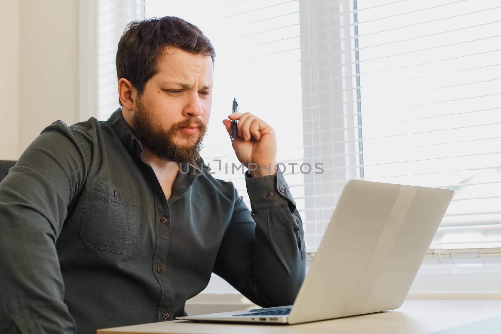 Caucasian businessman sitting with pen and laptop at cabinet. Concept of office worker and modern technology. Adult man wearing shirt and surfing internet.