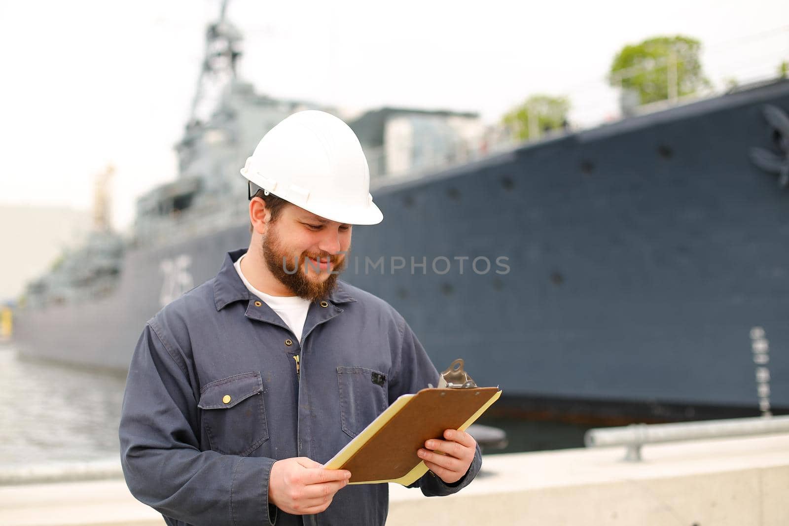 Deckhand wearing helmet and holding papers documents, standing on coast near vessel. Concept of maritime job and engineering department of marine team.