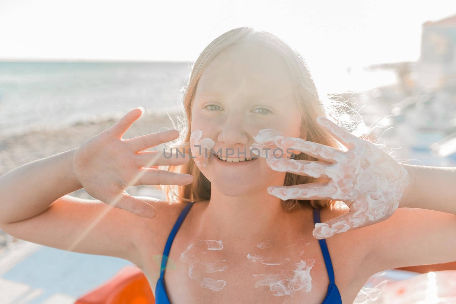 Portrait of a young positive teenage girl blonde of European appearance with sunscreen on her face and body against the background of a sea beach.