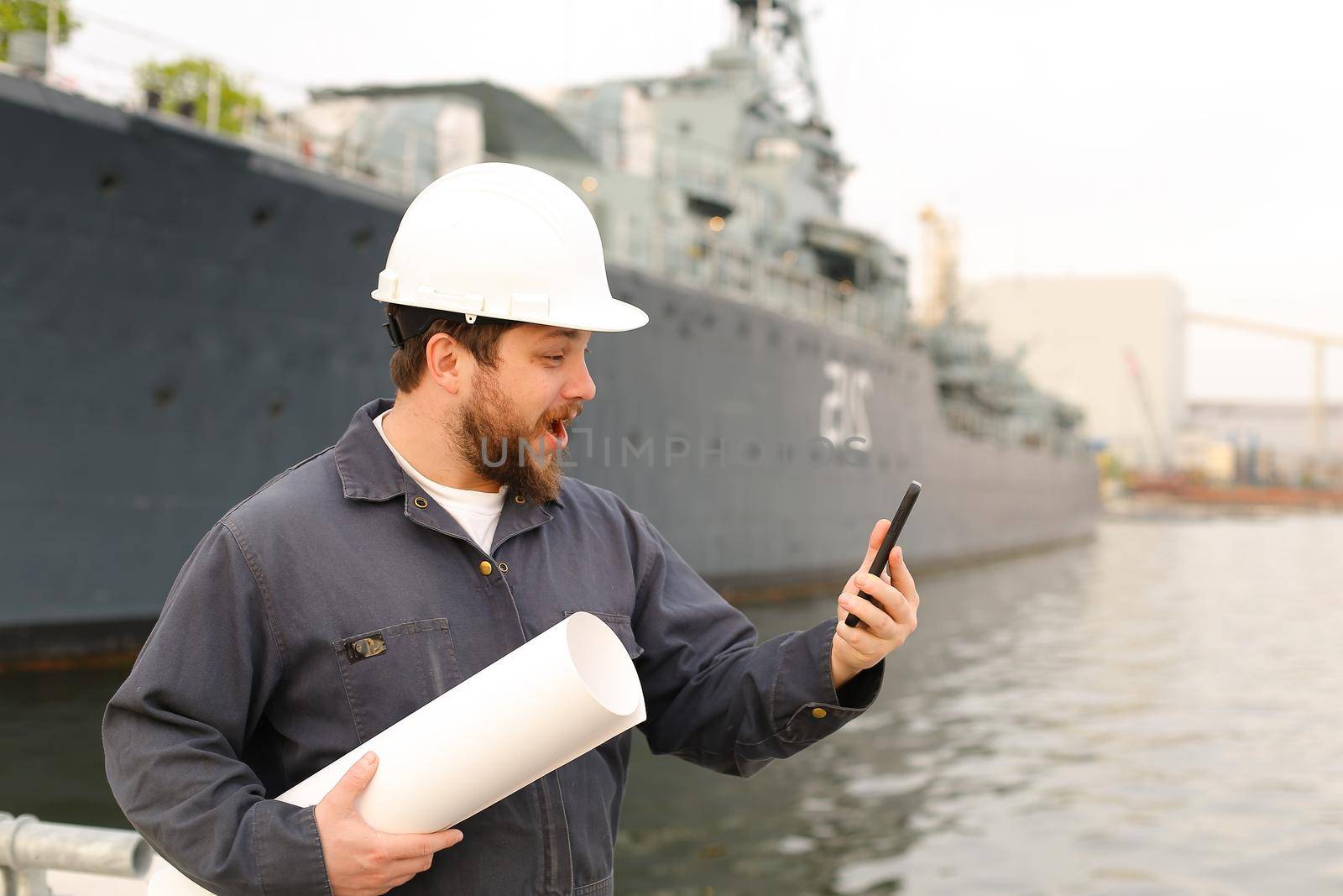 Marine engineer holding VHF walkie talkie and blueprints near vessel in background, wearing helmet and work jumpsuit. Concept of maritime profession , job and seaman.