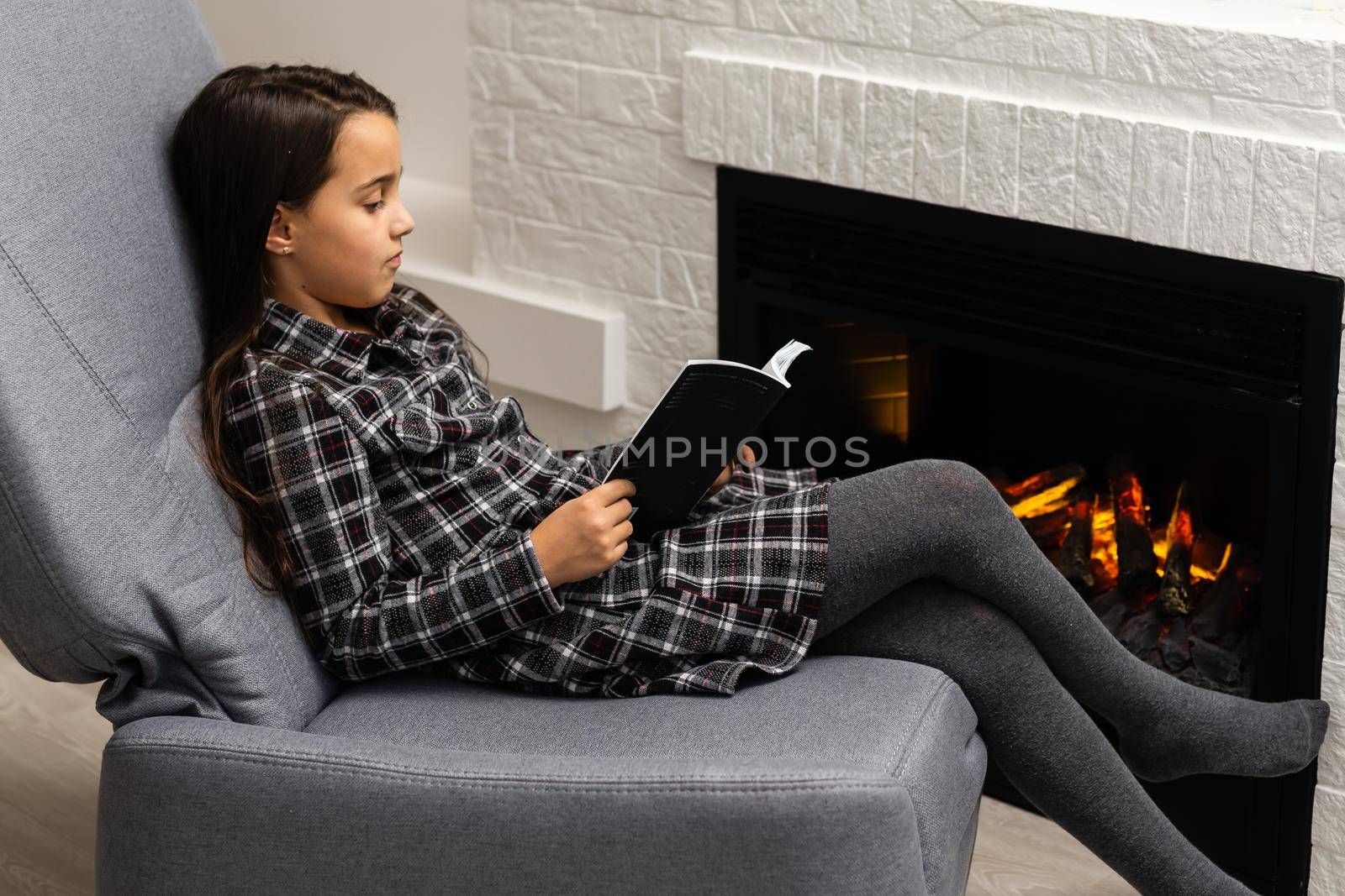 Little girl in dress sits in chair with book near fireplace