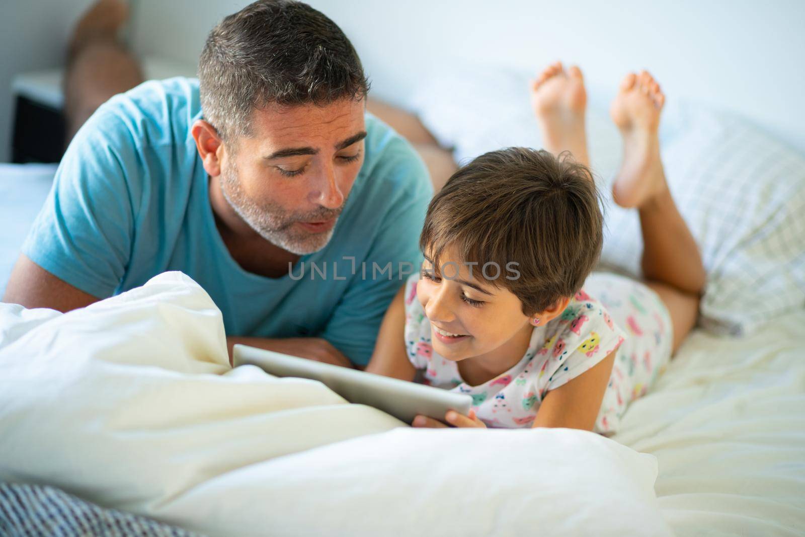 Funny dad and lovely little girl are having fun on the bed. Middle-age father with her eight years daughter using digital tablet in bedroom.