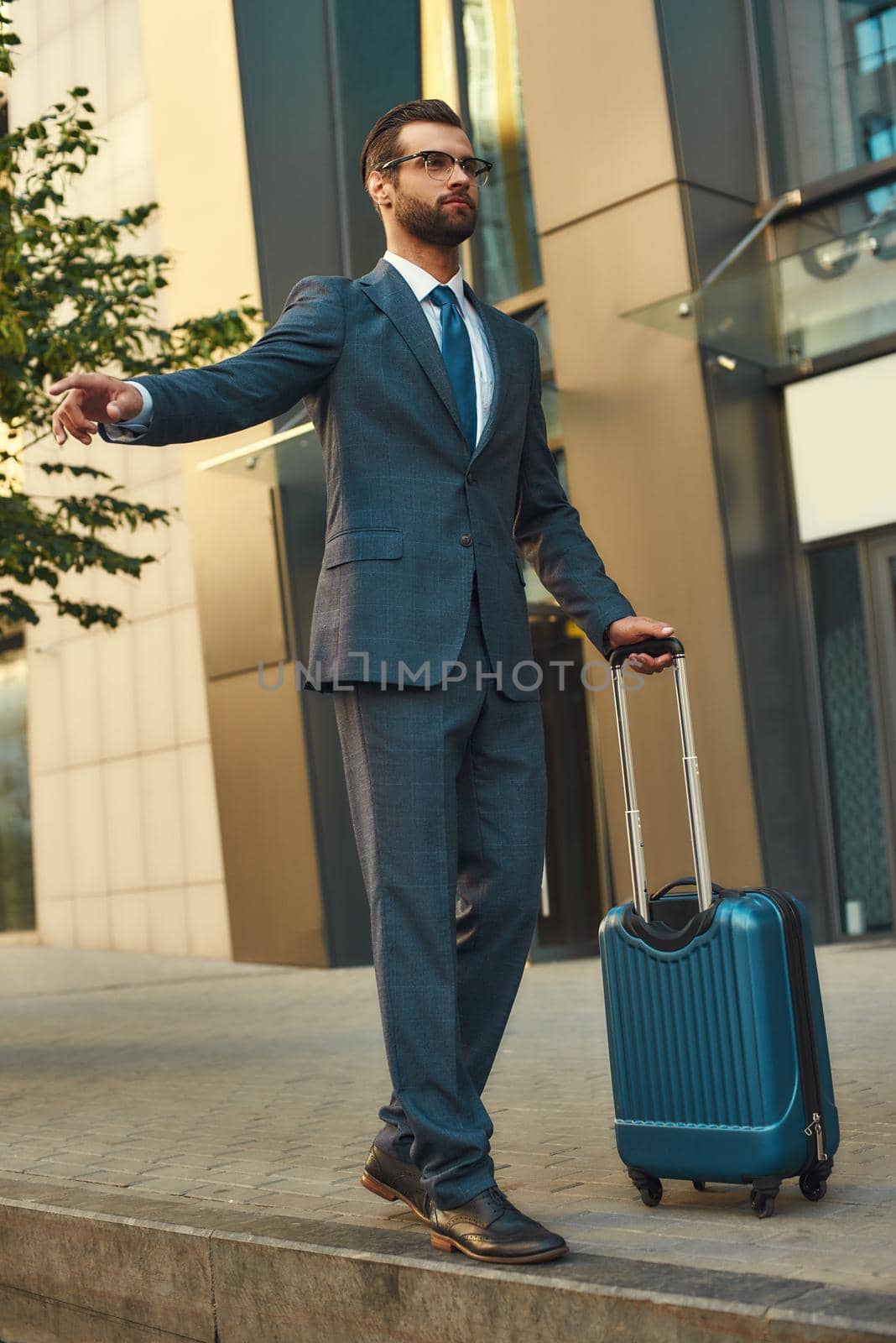 Catching taxi. Full length of young and handsome bearded man in suit carrying suitcase and raising his arm while standing outdoors by friendsstock