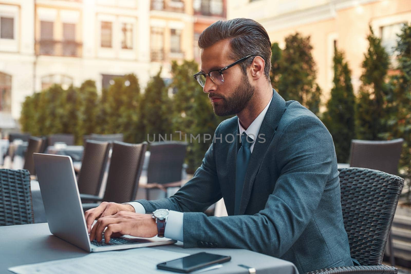 Modern businessman. Portrait of handsome bearded businessman in eyeglasses working with laptop while sitting in restaurant outdoors by friendsstock