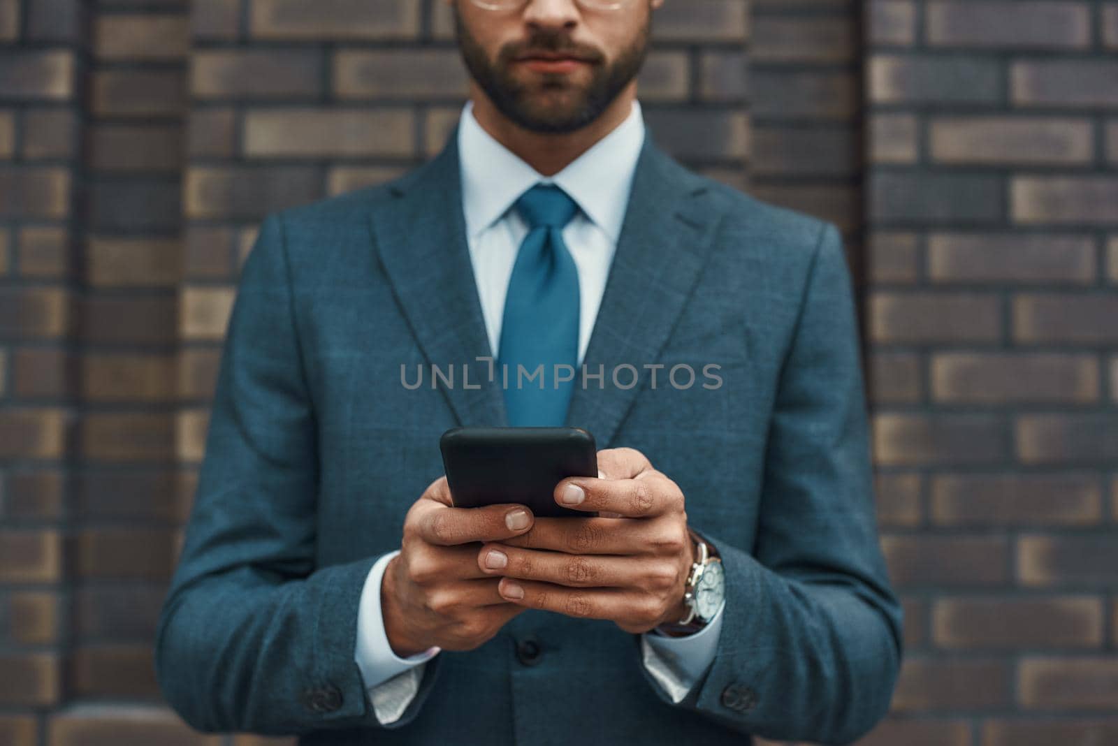 Cropped photo of a businessman in formal wear and using his smart phone while standing against brick wall. Business concept. Digital concept