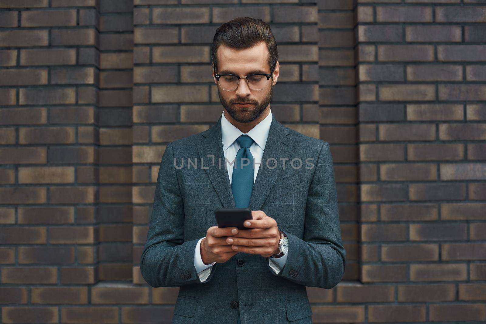 Business message. Handsome businessman in formal wear and eyeglasses using smart phone while standing against brick wall. Business concept. Digital concept