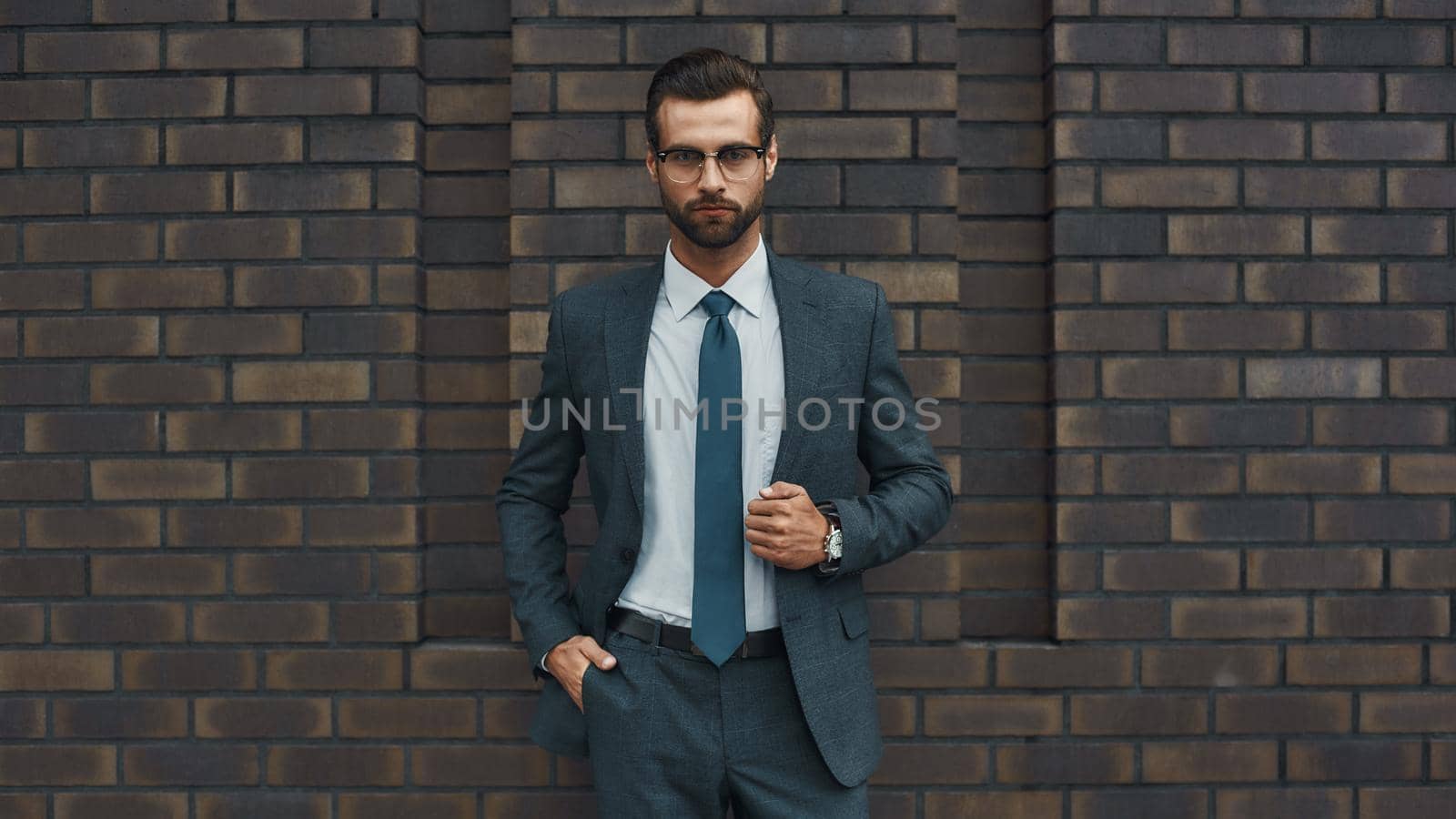 Great style. Confident businessman in full suit keeping hand in pocket and looking at camera while standing against brick wall. Business concept. Stylish people