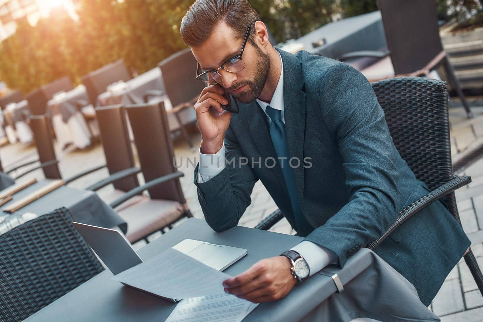 Working from everywhere. Portrait of handsome bearded businessman in eyeglasses talking by phone with client and looking at documents while sitting in restaurant outdoors by friendsstock