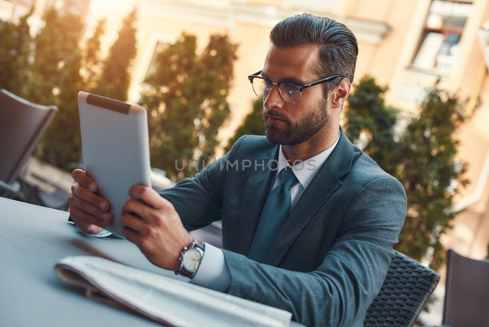 Modern businessman. Portrait of handsome bearded businessman in eyeglasses working with touchpad while sitting in restaurant outdoors by friendsstock