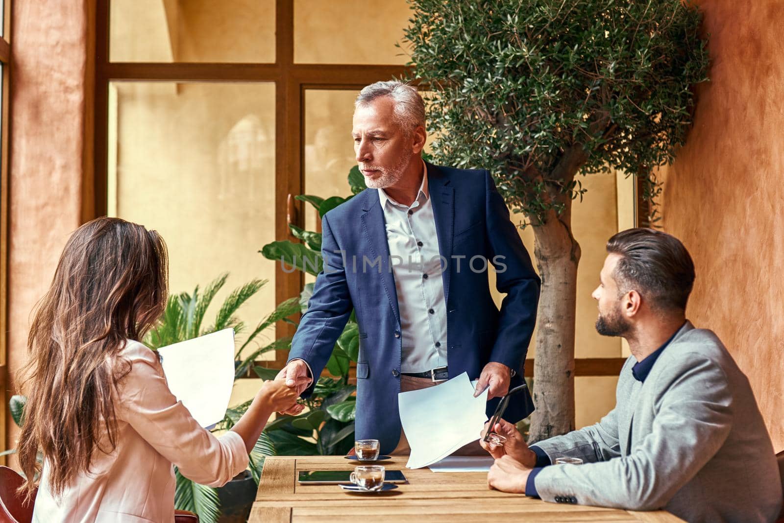 Business lunch. Three people in the restaurant sitting at table making deal senior man shaking hands with young woman. Agreement concept by friendsstock