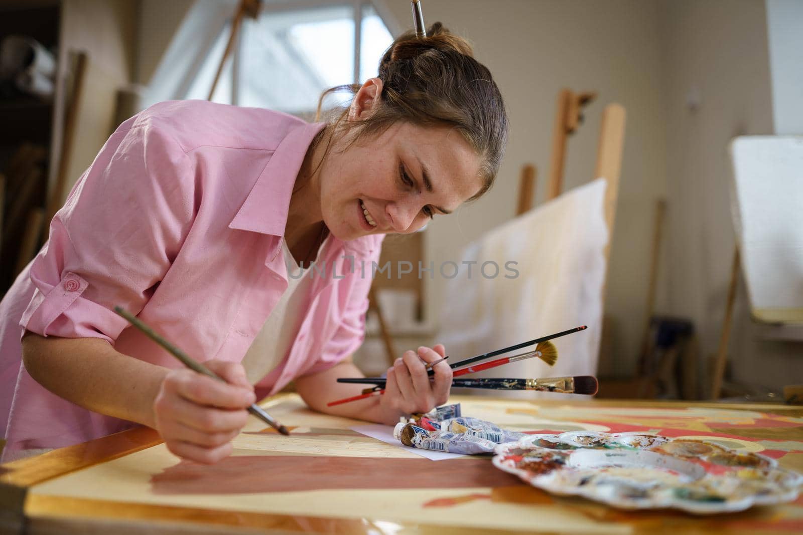 Caucasian woman artist working on a painting in bright daylight studio. Happy artist draws an art project with paints and a brush in the workshop. Hobby. Artist at work. Creative profession.